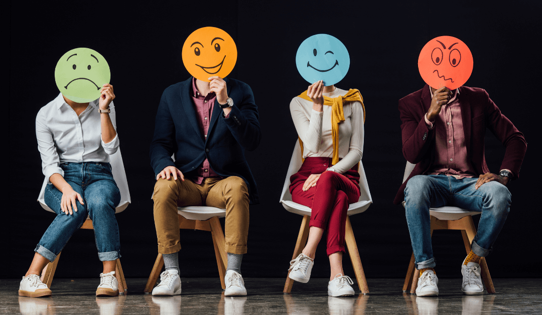 Four people sitting on chairs. Each holds a smily face with a different emotional expression in front of their face.