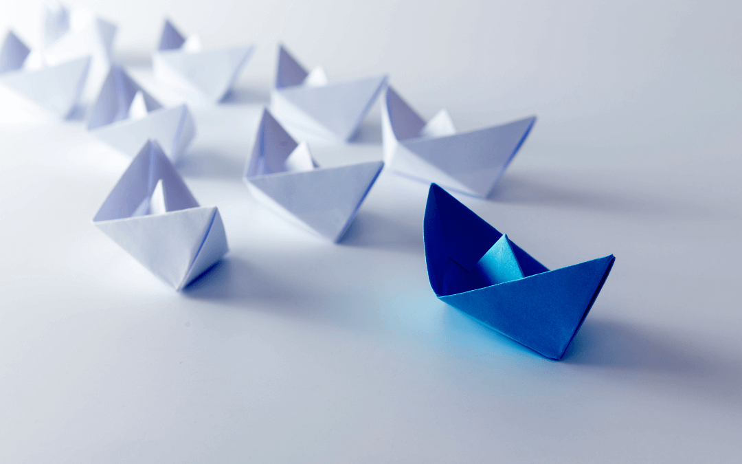 A blue paper boat ahead of white paper boats. Symbolises leadership.