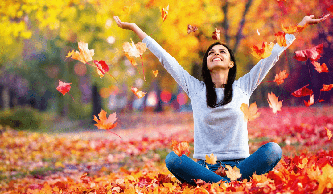 Woman sitting in pile of fallen leaves. She smiles with joy.